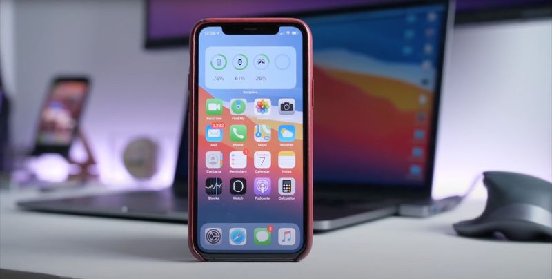 A photograph showing an iPhone in the foreground with the iOS 14 Home Screen and a MacBook Pro in the background