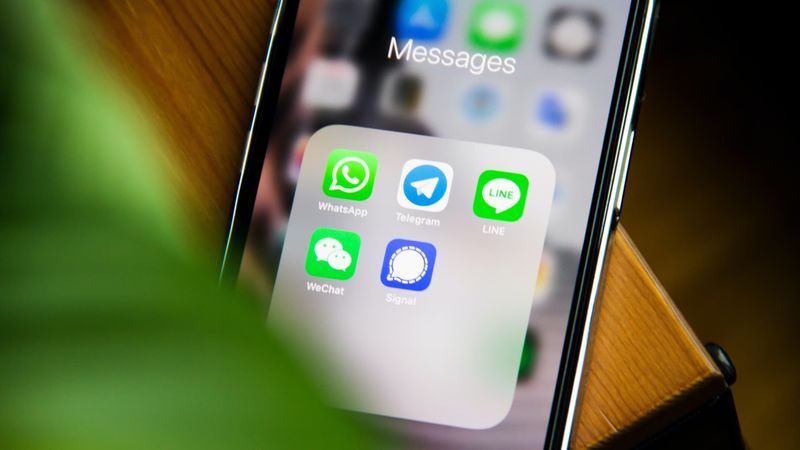 A photograph of an iPhone laid back on a table edge, with the Messages apps folder showing icons for WhatsApp, Telegram, LINE, WeChat and Signal