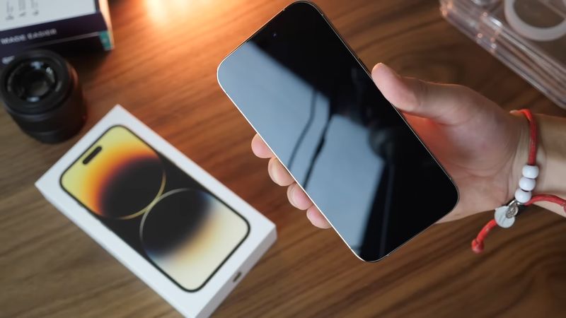 Male hand holding iPhone 14 Pro Max with its alway-on display completely dark and product packaging visible in the background