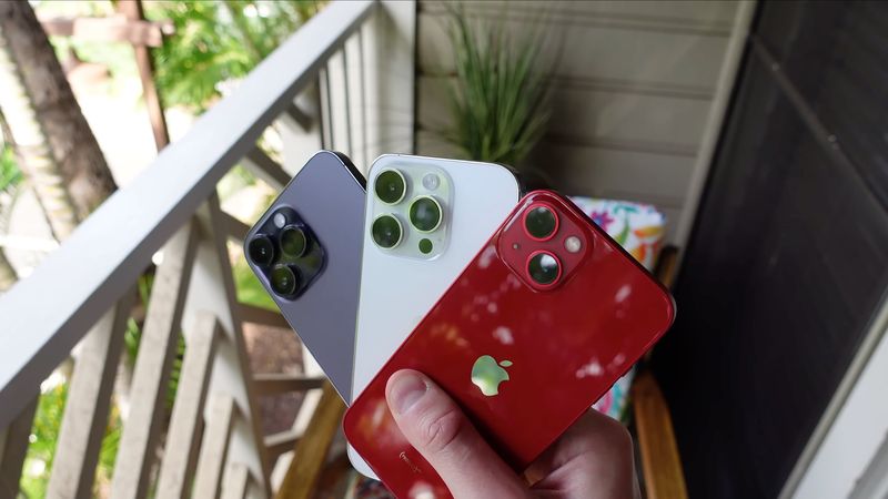 Young male hand holding purple and silver iPhone 14 Pros and a red iPhone 14, with a wooden back porch in the background