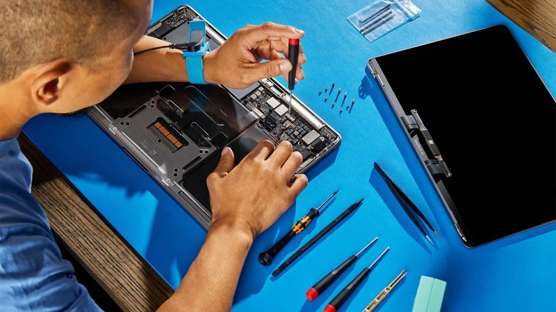 A customer hunched over their table, using genuine Apple parts, tools and manuals to repair a broken MacBook Pro