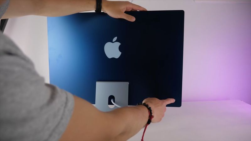 A photograph showing the backside of a blue 24-inch Apple M1 iMac computer with a finger touching the power button