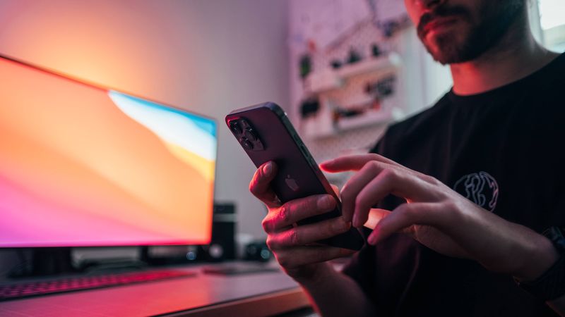 Young man holding an iPhone 12 Pro Max in front of his work desk. In the background, there