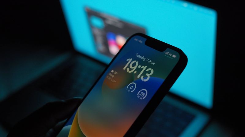 iPhone 13 Pro Max displaying an iOS 16 Lock Screen, held in hand above a MacBook Air