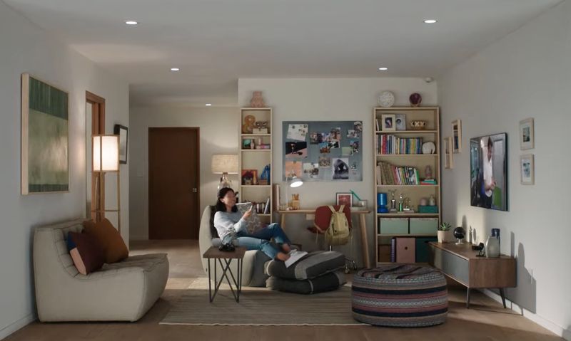 Une image de style de vie montrant une maison avec une jeune femme assise sur le canapé, souriant et regardant Apple TV et tenant Siri Remote dans sa main droite
